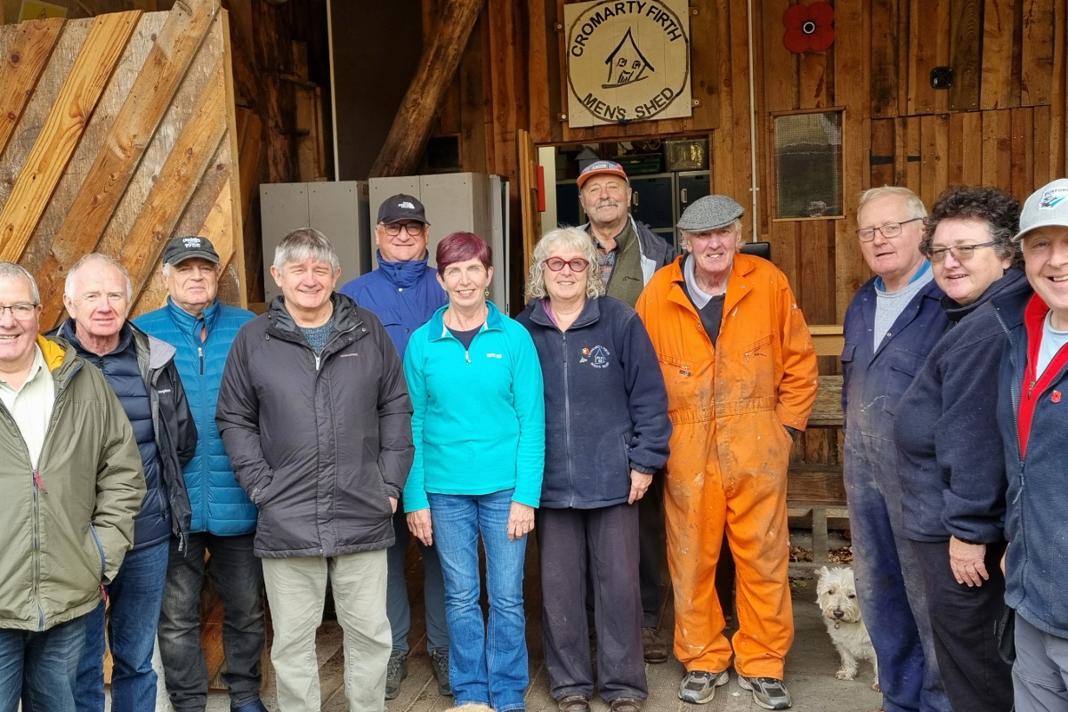 A group of people standing outside a large wooden cabin with a sign for Cromarty Firth men's Shed