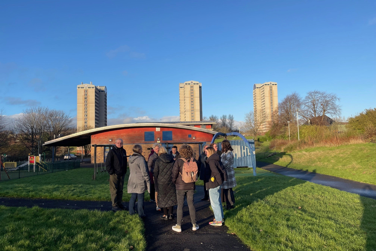 A group of people standing outside on a public walkway with a kids park and 3 high rise blocks in the distance.