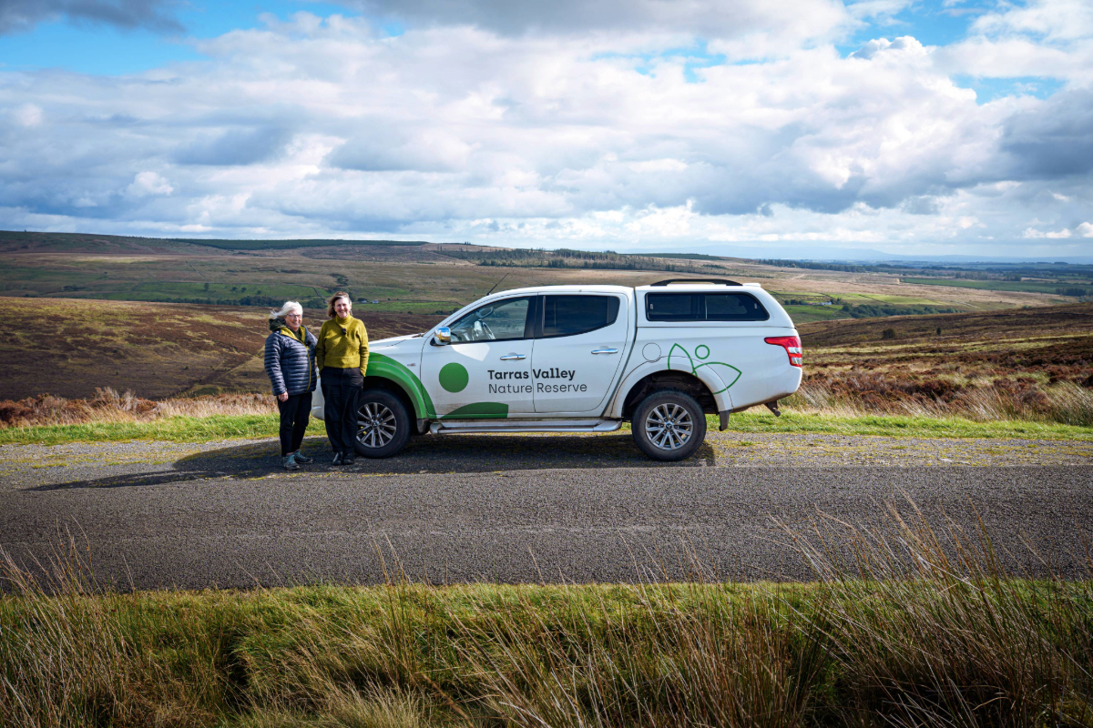 Two people standing in front of a white 4x4 ranger with the Tarras Valley nature reserve surrounding them