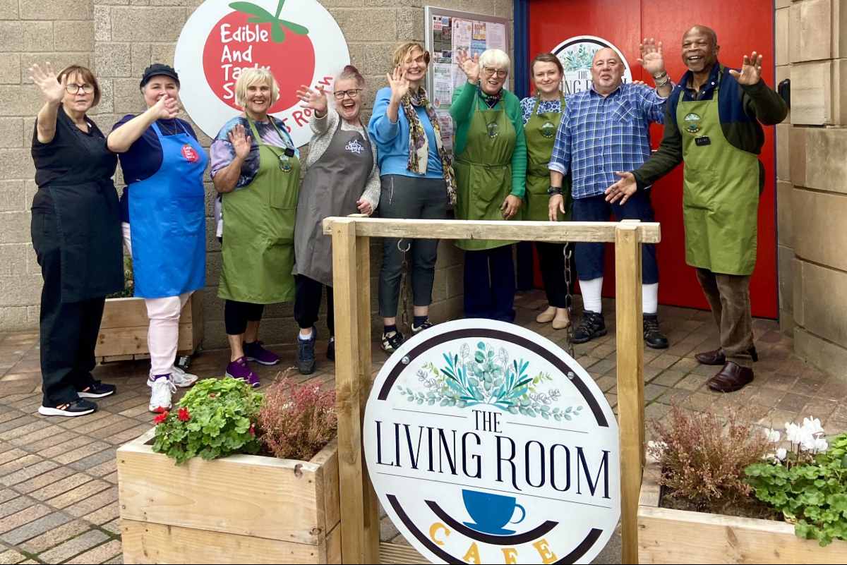 A group of 9 people smiling and waving to the camera behind planter boxes and a sign for 'The Livingroom' cafe