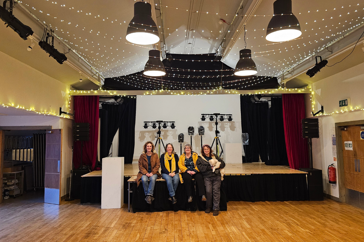 4 people sitting on a stage, one holding a small dog in a community hall with fairy lights suspended from the ceiling