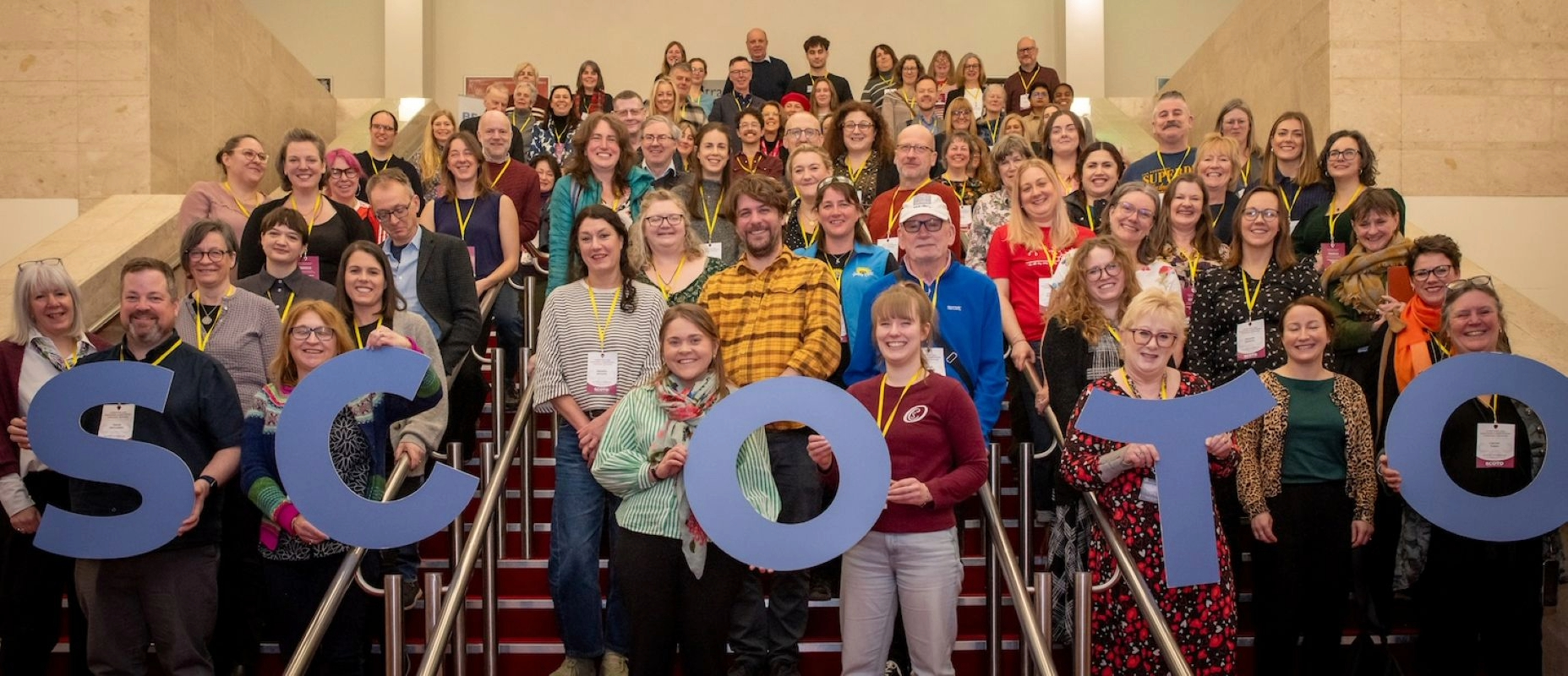 A large group of people standing on a stairway with people at the front holding large letters spelling out SCOTO