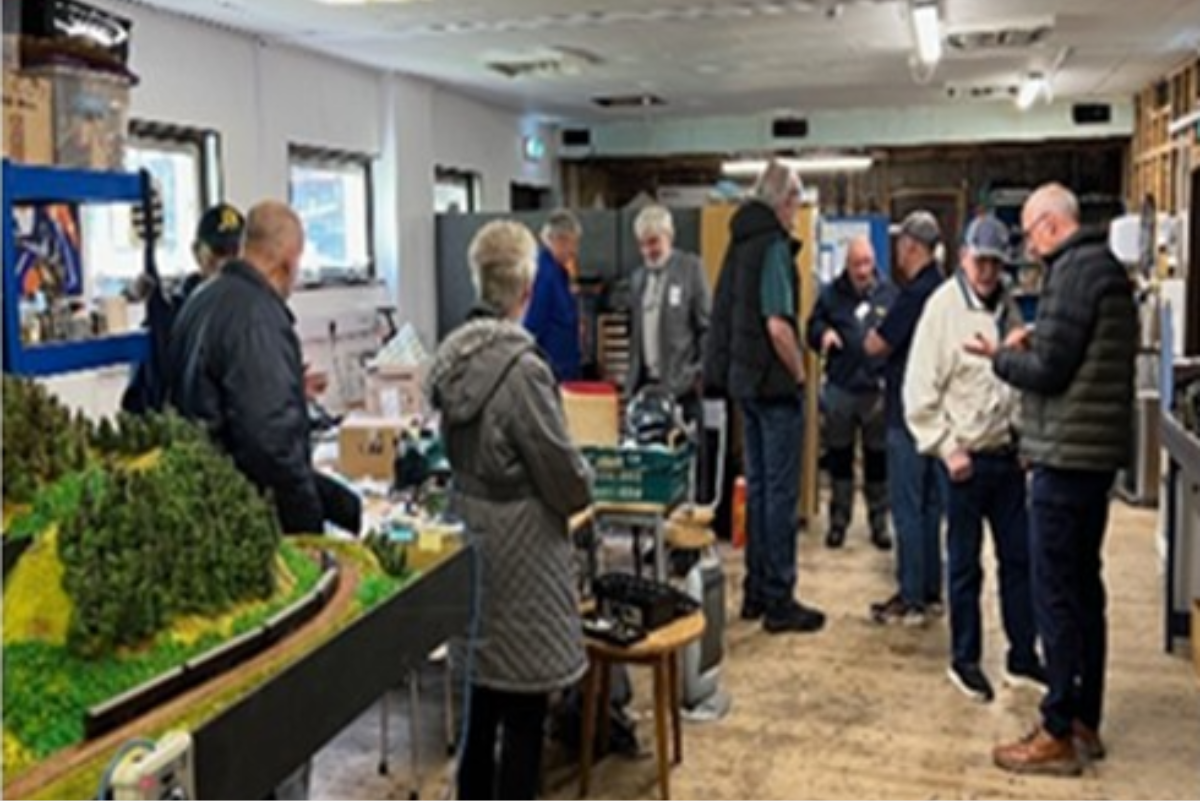 A group of 10 people viewing a workshop with a model railway, chairs and machinery dotted about the room