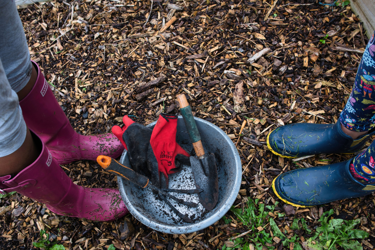 A bucket on the ground with a muddy trowel, garden fork and gloves inside. With a person legs and purple wellies on the left and a person blue wellies to the right of the bucket.